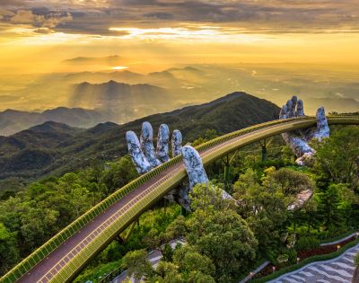 Golden bridge with giant stone hands emerges from lush forest, set against a glowing sunset over distant mountains and valleys, conveying awe and serenity.