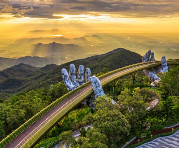 Golden bridge with giant stone hands emerges from lush forest, set against a glowing sunset over distant mountains and valleys, conveying awe and serenity.
