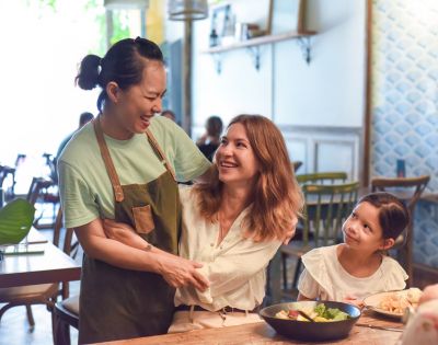 Two women share a warm laugh in a cozy café, with one in an apron. A child smiles up at them from the table, creating a joyful, friendly atmosphere.