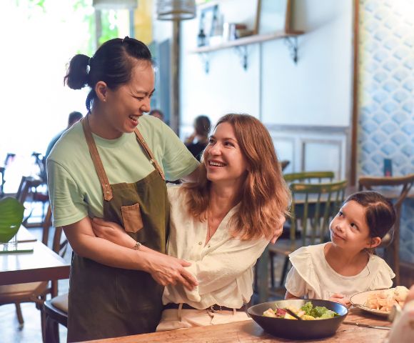 Two women share a warm laugh in a cozy café, with one in an apron. A child smiles up at them from the table, creating a joyful, friendly atmosphere.