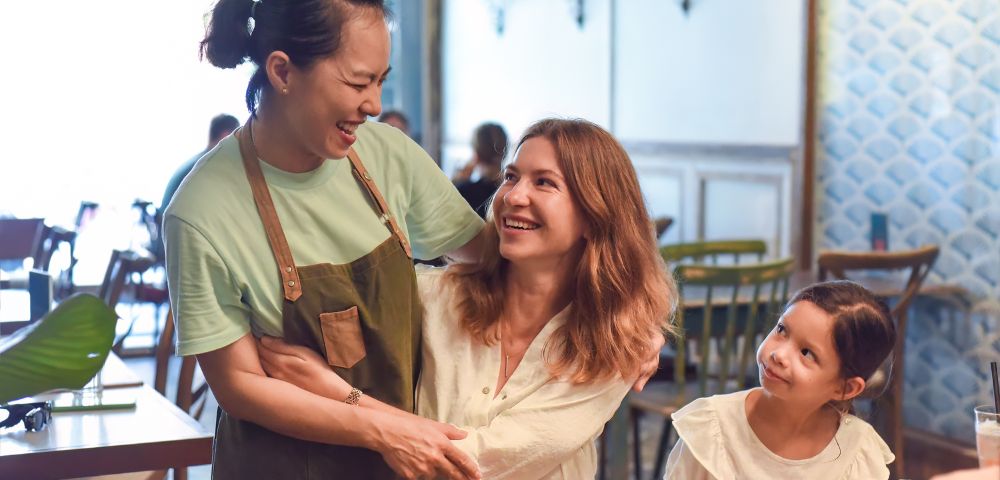 Two women share a warm laugh in a cozy café, with one in an apron. A child smiles up at them from the table, creating a joyful, friendly atmosphere.
