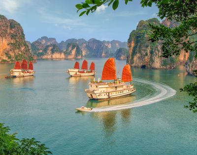 Four traditional boats with red sails glide through the tranquil waters of Halong Bay, surrounded by towering limestone cliffs under a clear blue sky.