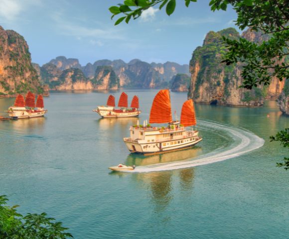 Four traditional boats with red sails glide through the tranquil waters of Halong Bay, surrounded by towering limestone cliffs under a clear blue sky.