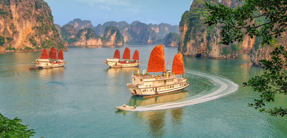Four traditional boats with red sails glide through the tranquil waters of Halong Bay, surrounded by towering limestone cliffs under a clear blue sky.