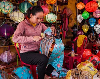 A woman in a pink hoodie assembles colorful lanterns in a vibrant shop filled with various lanterns, conveying a sense of focus and creativity.
