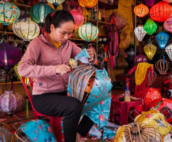 A woman in a pink hoodie assembles colorful lanterns in a vibrant shop filled with various lanterns, conveying a sense of focus and creativity.