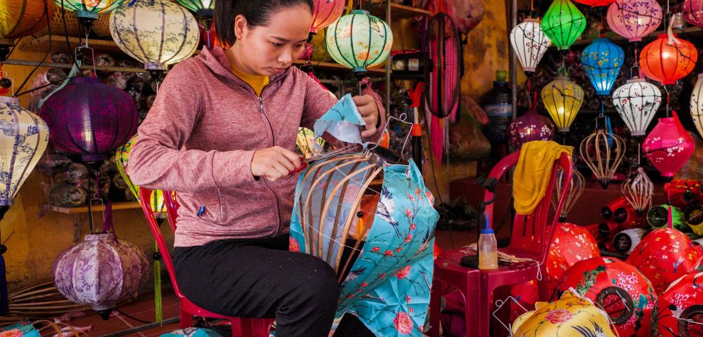 A woman in a pink hoodie assembles colorful lanterns in a vibrant shop filled with various lanterns, conveying a sense of focus and creativity.