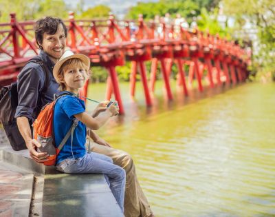 Father and son, smiling, sit by a serene lake with a vivid red wooden bridge in the background. The scene is sunny and joyful, evoking a sense of adventure.