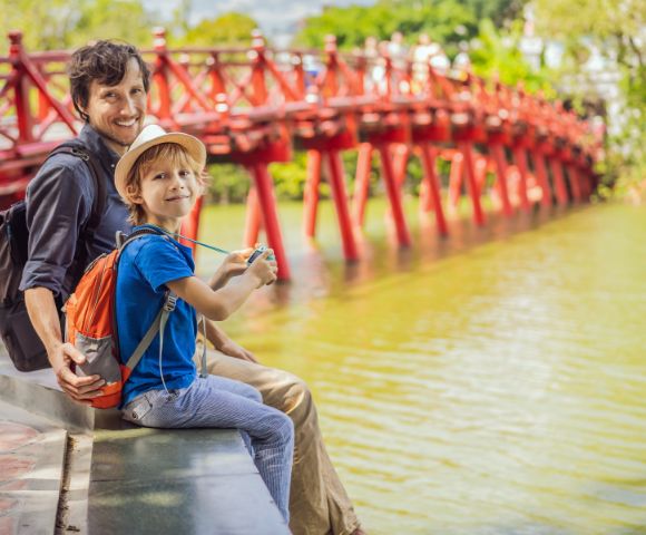 Father and son, smiling, sit by a serene lake with a vivid red wooden bridge in the background. The scene is sunny and joyful, evoking a sense of adventure.