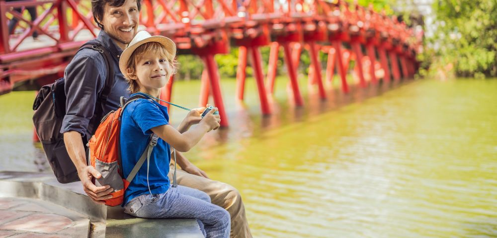 Father and son, smiling, sit by a serene lake with a vivid red wooden bridge in the background. The scene is sunny and joyful, evoking a sense of adventure.