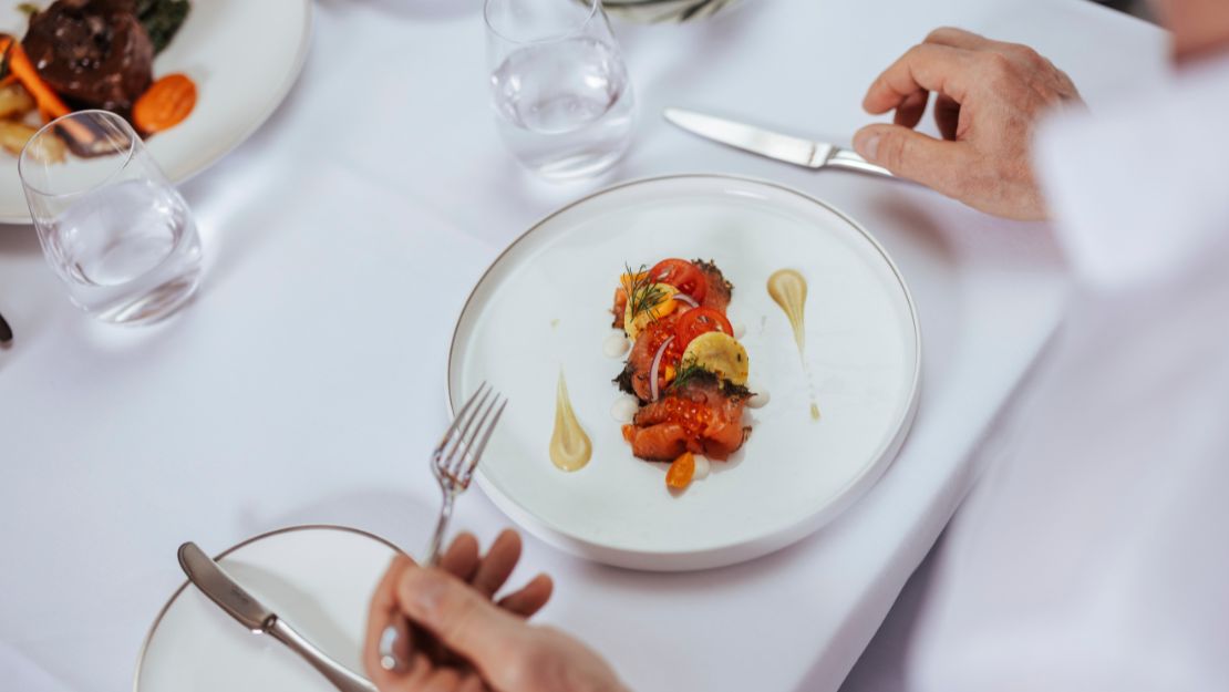Elegant dining setting with a gourmet dish on a white plate featuring colorful vegetables, artfully drizzled sauce, and hands holding cutlery, conveying a refined atmosphere.