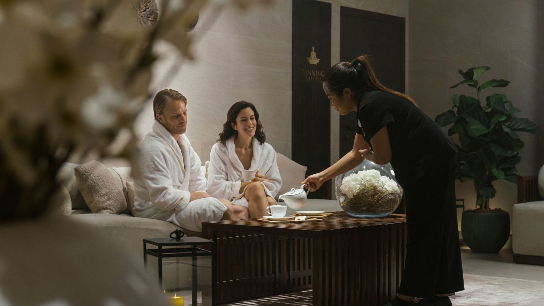 A couple in white robes sit on a cozy sofa in a serene spa waiting area. A staff member pours tea from a teapot on a wooden table, creating a relaxing ambiance.