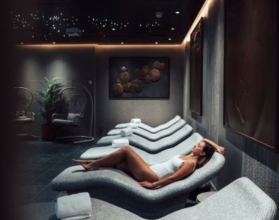 A woman in a white swimsuit relaxes on a curved lounge chair in a dimly lit spa room. The ambience is serene, with soft lighting and modern decor.