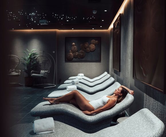 A woman in a white swimsuit relaxes on a curved lounge chair in a dimly lit spa room. The ambience is serene, with soft lighting and modern decor.