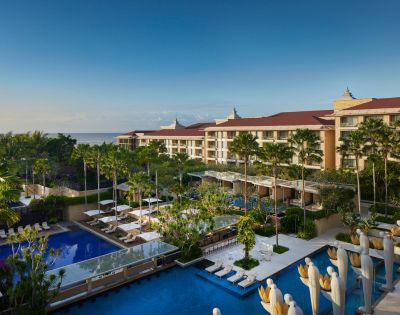 Luxurious resort overlooking the ocean, with multiple swimming pools surrounded by palm trees and sunbeds. The sky is clear, conveying a serene atmosphere.