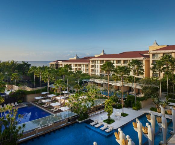 Luxurious resort overlooking the ocean, with multiple swimming pools surrounded by palm trees and sunbeds. The sky is clear, conveying a serene atmosphere.