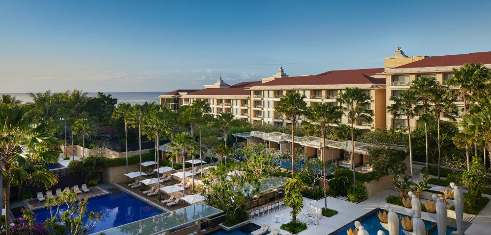 Luxurious resort overlooking the ocean, with multiple swimming pools surrounded by palm trees and sunbeds. The sky is clear, conveying a serene atmosphere.