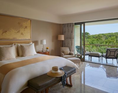 A serene hotel room with a king-sized bed, soft lighting, and a balcony offering a lush green view. Cozy seating includes an armchair and outdoor chairs.