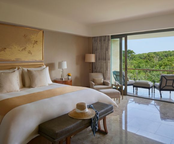 A serene hotel room with a king-sized bed, soft lighting, and a balcony offering a lush green view. Cozy seating includes an armchair and outdoor chairs.