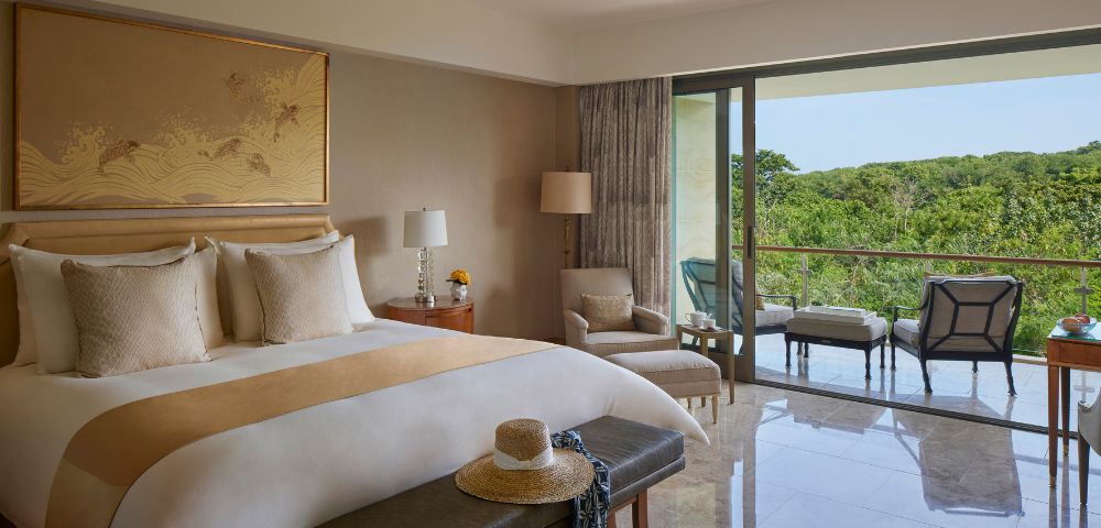 A serene hotel room with a king-sized bed, soft lighting, and a balcony offering a lush green view. Cozy seating includes an armchair and outdoor chairs.