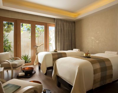 A serene spa room with two massage tables adorned with beige blankets. Soft lighting, wooden floor, and large windows looking out to greenery create a tranquil atmosphere.