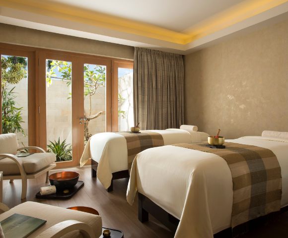 A serene spa room with two massage tables adorned with beige blankets. Soft lighting, wooden floor, and large windows looking out to greenery create a tranquil atmosphere.