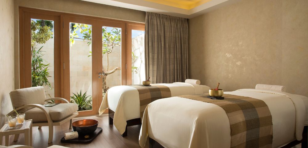 A serene spa room with two massage tables adorned with beige blankets. Soft lighting, wooden floor, and large windows looking out to greenery create a tranquil atmosphere.