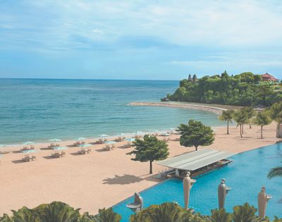 A serene beach scene with turquoise waters and a sandy shore lined with white lounge chairs and umbrellas. Lush greenery and a tranquil pool enhance the coastal vibe.