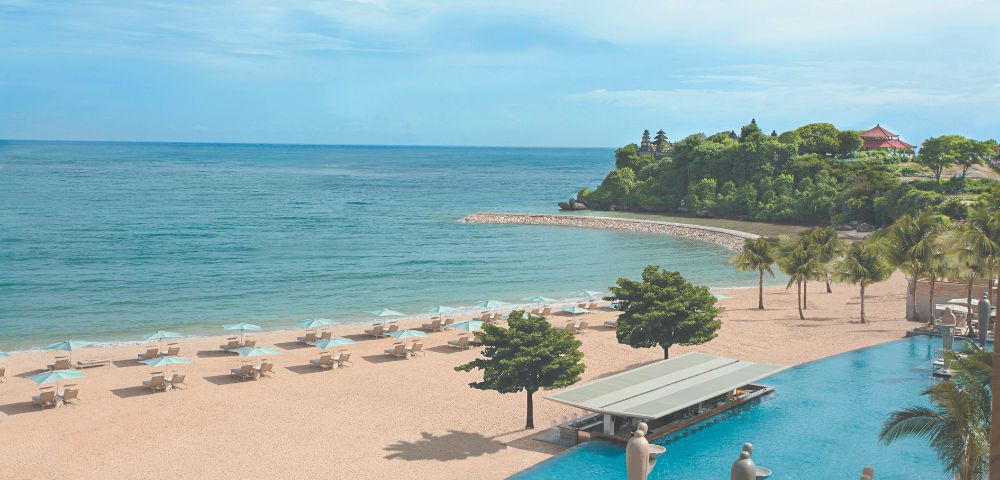 A serene beach scene with turquoise waters and a sandy shore lined with white lounge chairs and umbrellas. Lush greenery and a tranquil pool enhance the coastal vibe.