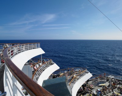 A ship deck with curved railings overlooks a vast ocean under a clear blue sky. Sunbathers relax in lounge chairs, conveying a peaceful, leisurely atmosphere.