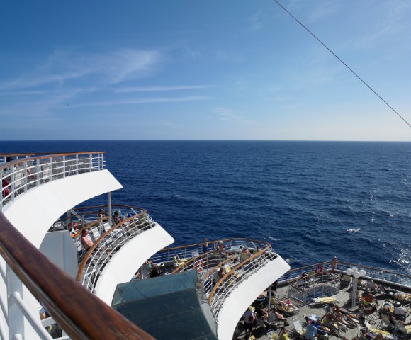 A ship deck with curved railings overlooks a vast ocean under a clear blue sky. Sunbathers relax in lounge chairs, conveying a peaceful, leisurely atmosphere.