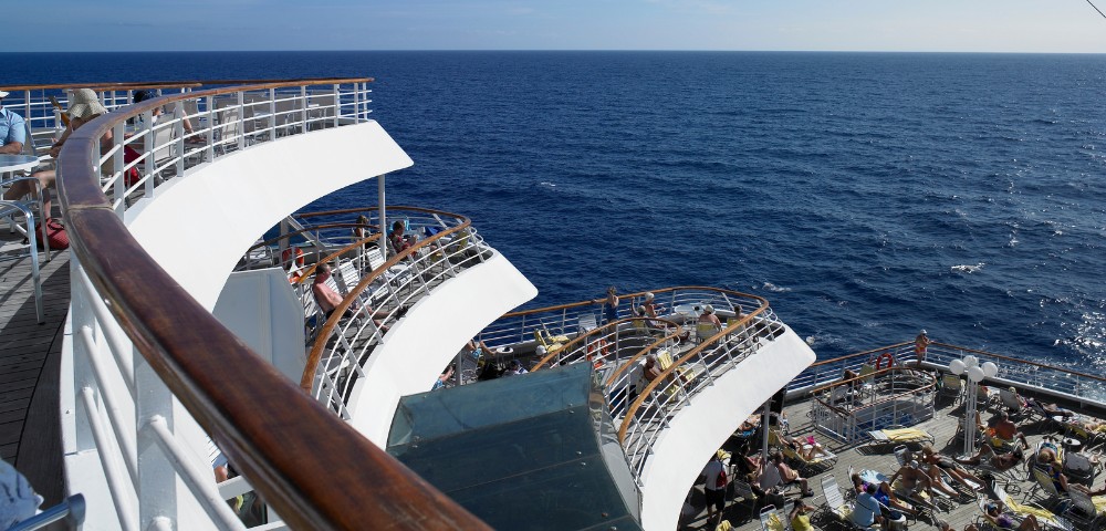 A ship deck with curved railings overlooks a vast ocean under a clear blue sky. Sunbathers relax in lounge chairs, conveying a peaceful, leisurely atmosphere.