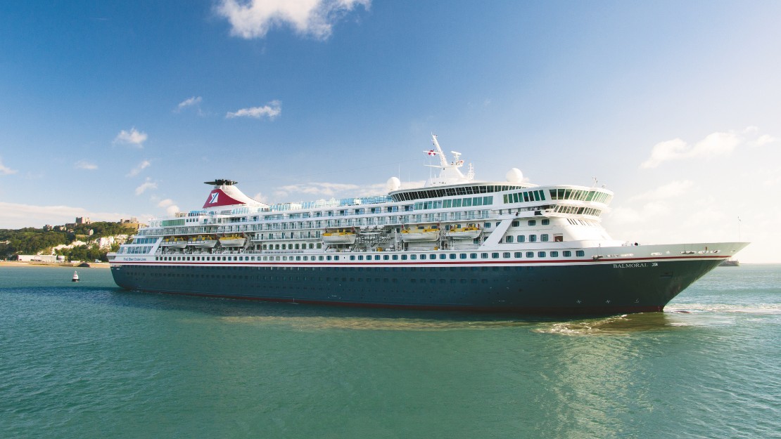 A large cruise ship sails in a calm, blue sea under a clear sky with a few clouds. The ship is white with a red funnel, conveying a sense of tranquility.