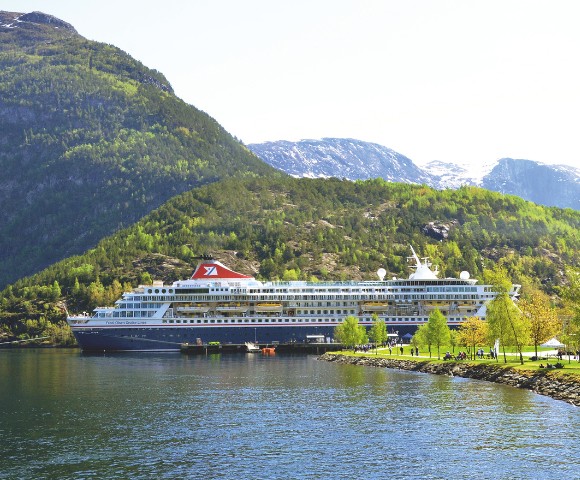 Cruise ship docked in a serene fjord, surrounded by lush green hills and rocky mountains under a bright, clear sky. Calm water reflects the vibrant landscape.