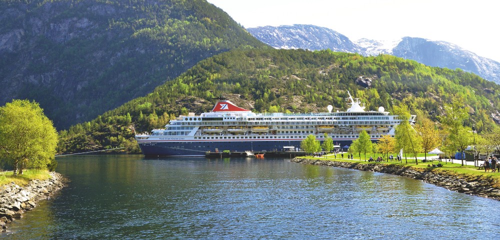 Cruise ship docked in a serene fjord, surrounded by lush green hills and rocky mountains under a bright, clear sky. Calm water reflects the vibrant landscape.