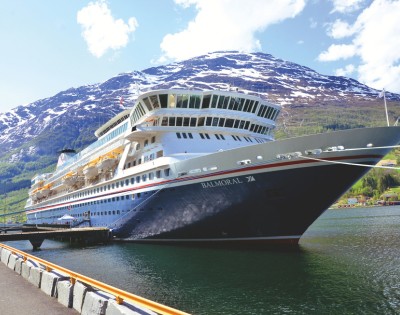Cruise ship docked in a scenic fjord, with snow-capped mountains and lush green hills in the background under a partly cloudy sky.