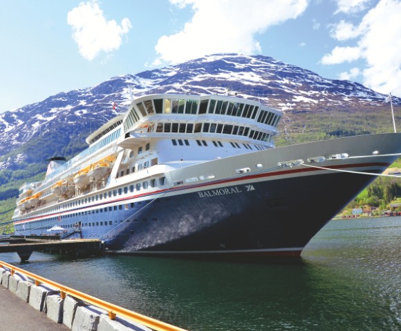 Cruise ship docked in a scenic fjord, with snow-capped mountains and lush green hills in the background under a partly cloudy sky.