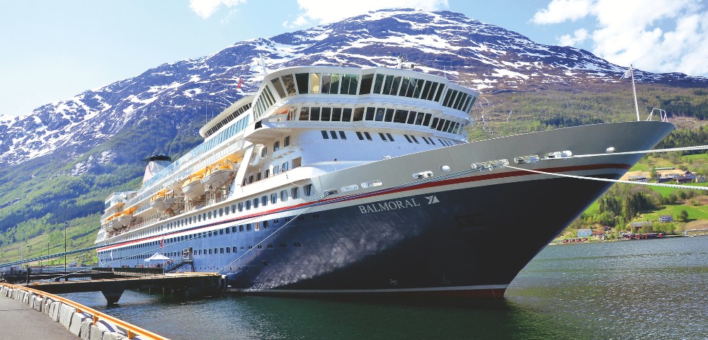 Cruise ship docked in a scenic fjord, with snow-capped mountains and lush green hills in the background under a partly cloudy sky.