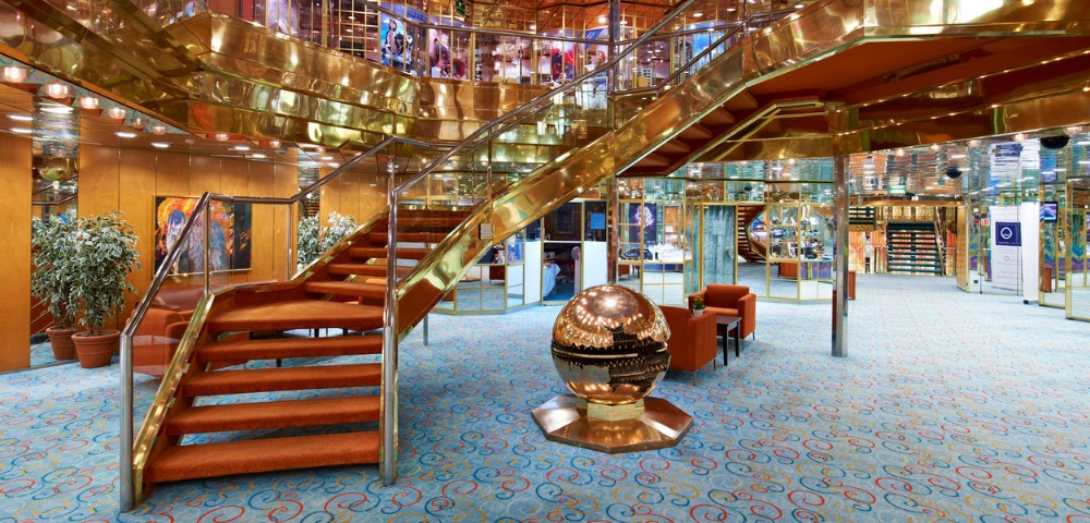 Elegant cruise ship lobby with grand staircase, plush orange carpet, and golden railings. Reflective globe sculpture in the center. Luxurious and inviting ambiance.
