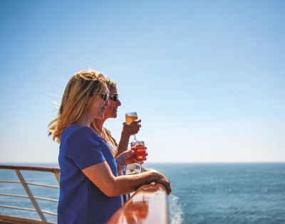 Two women stand on a cruise ship deck, enjoying drinks and gazing at the ocean under a clear blue sky. They appear relaxed and content.