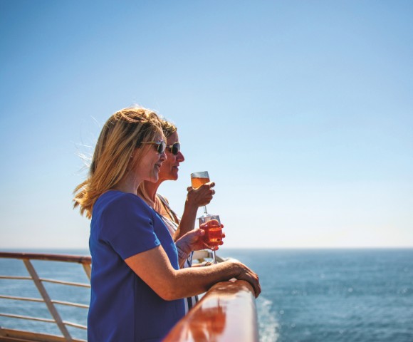 Two women stand on a cruise ship deck, enjoying drinks and gazing at the ocean under a clear blue sky. They appear relaxed and content.