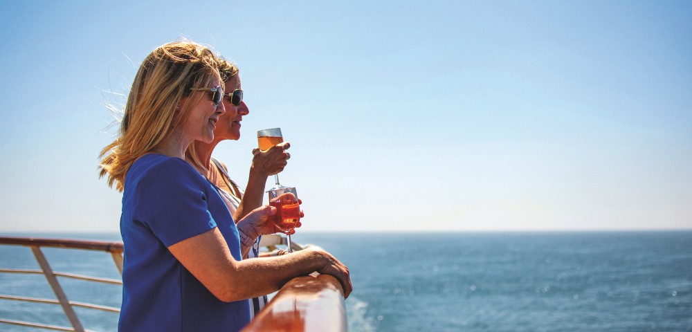 Two women stand on a cruise ship deck, enjoying drinks and gazing at the ocean under a clear blue sky. They appear relaxed and content.