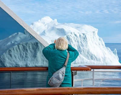A person in a teal sweater is on a ship, photographing a large iceberg nearby. The scene conveys awe and wonder against a bright, clear sky.