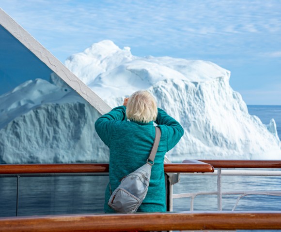 A person in a teal sweater is on a ship, photographing a large iceberg nearby. The scene conveys awe and wonder against a bright, clear sky.