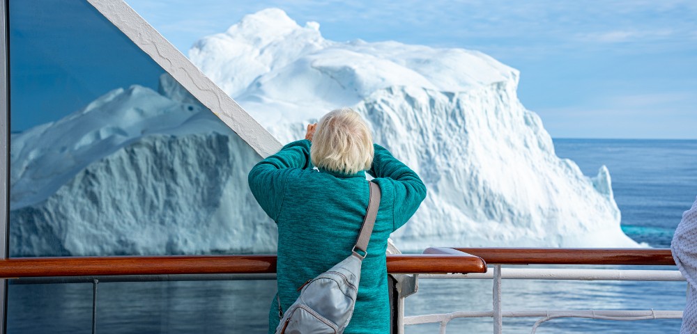 A person in a teal sweater is on a ship, photographing a large iceberg nearby. The scene conveys awe and wonder against a bright, clear sky.