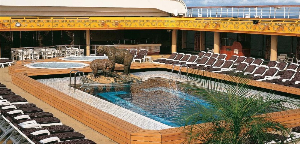 Rooftop pool area on a cruise ship features two jacuzzis, bear sculptures in the center, surrounded by lounge chairs. Sunny and inviting atmosphere.