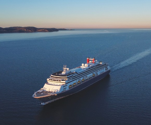 A large cruise ship sails through calm, expansive blue waters under a clear sky at sunset, with distant hills on the horizon, conveying tranquility.