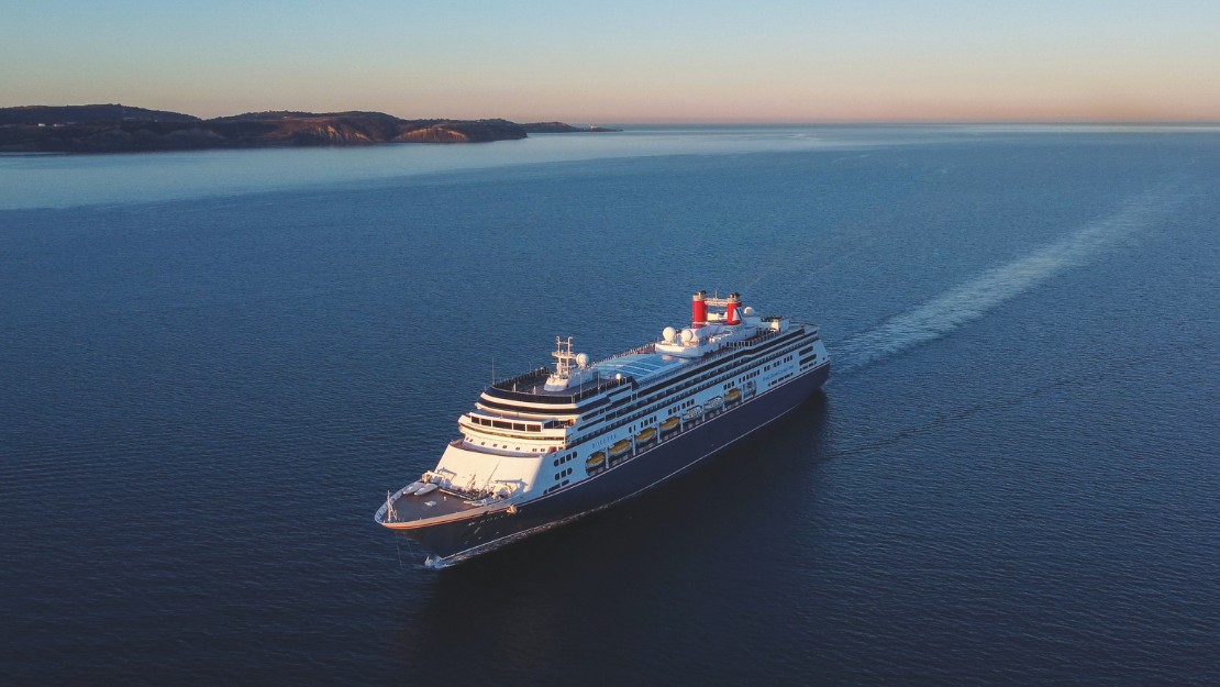 A large cruise ship sails through calm, expansive blue waters under a clear sky at sunset, with distant hills on the horizon, conveying tranquility.