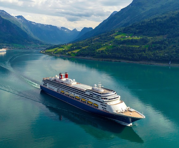A large cruise ship sails through a calm fjord, surrounded by lush green hills and distant snow-capped mountains under a bright sky. Tranquil and majestic.