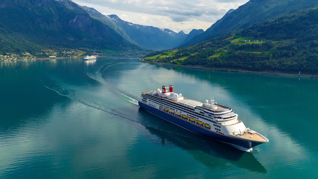 A large cruise ship sails through a calm fjord, surrounded by lush green hills and distant snow-capped mountains under a bright sky. Tranquil and majestic.
